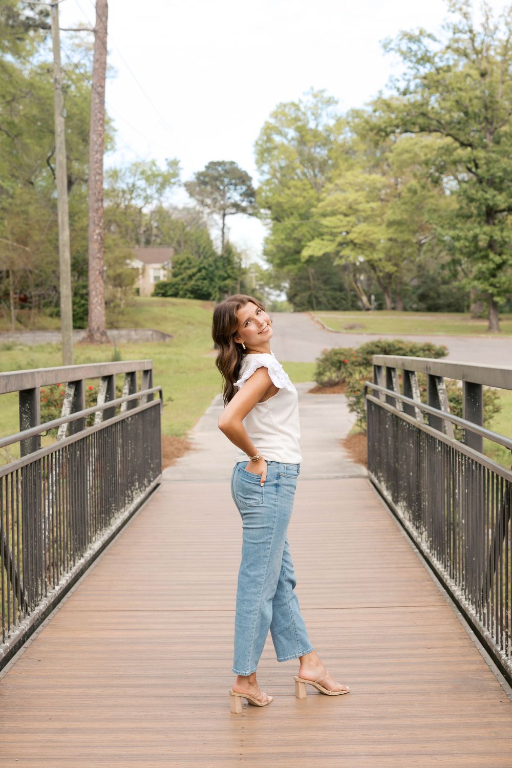 Young woman in white top and blue jeans posing on a wooden bridge surrounded by lush green trees.