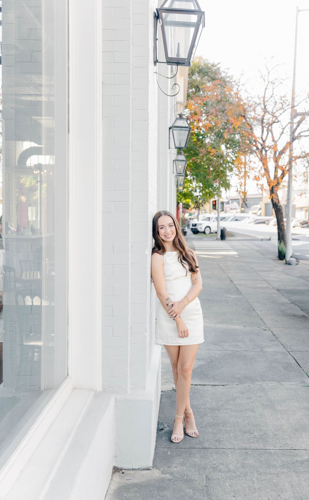 Young woman in white dress posing on urban sidewalk beside vintage gas lanterns and white building facade.