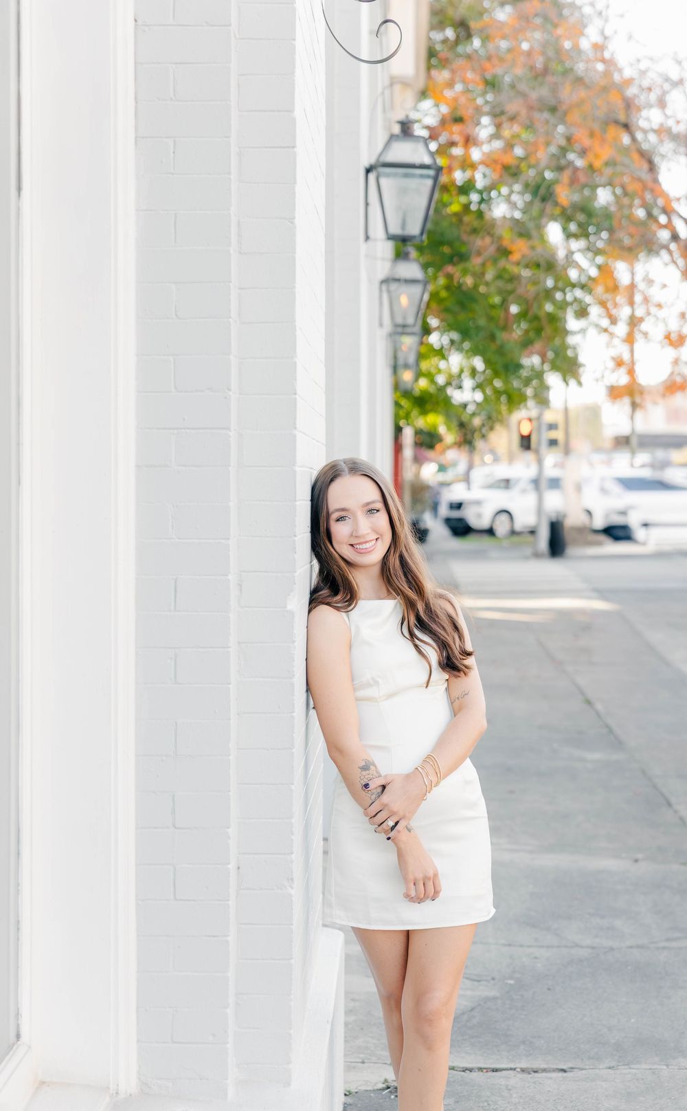 Young woman in white mini dress smiling beside brick wall with gas lamp on sunny sidewalk.