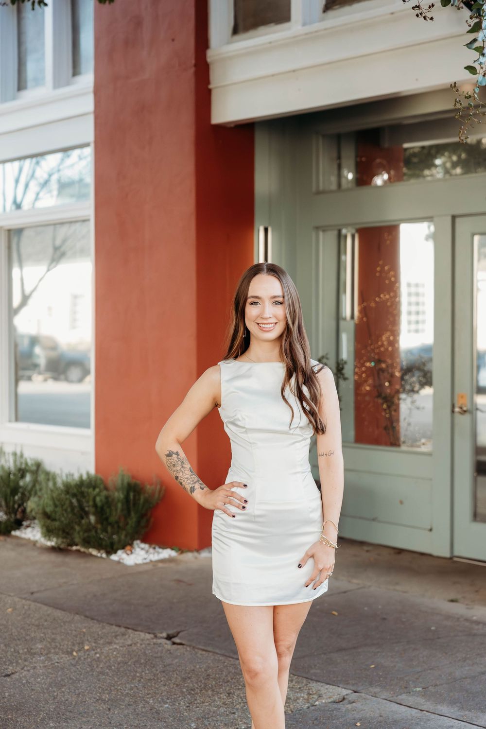 Young woman in white mini dress posing confidently on sidewalk outside building with green doors.