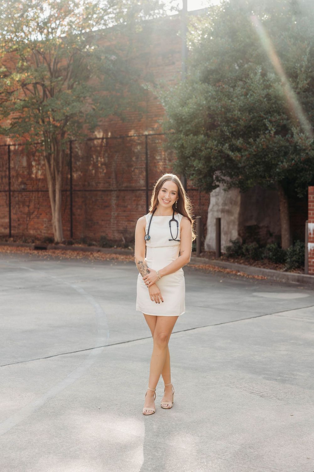 Young woman in white mini dress with stethoscope poses outdoors near brick building and green trees.