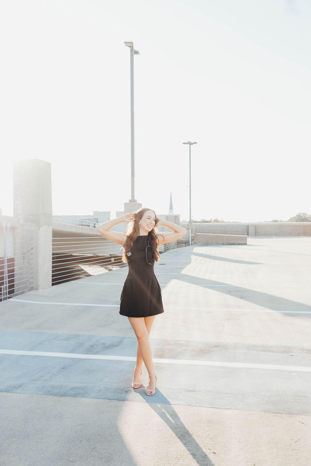 Young woman in black mini dress posing on rooftop parking deck in bright sunlight.
