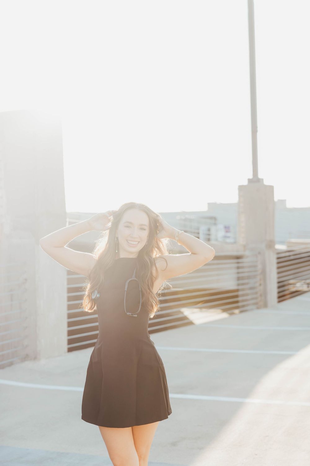 Young woman in black dress smiling on rooftop with sun flare backdrop, hands in hair.