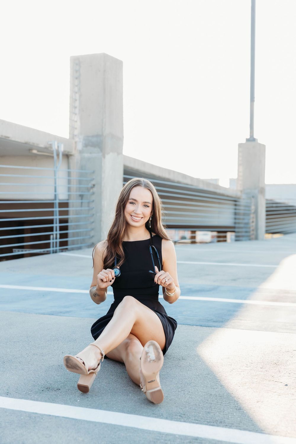 Young woman in black dress sitting on rooftop parking lot smiling, holding sunglasses, wearing nude heels.