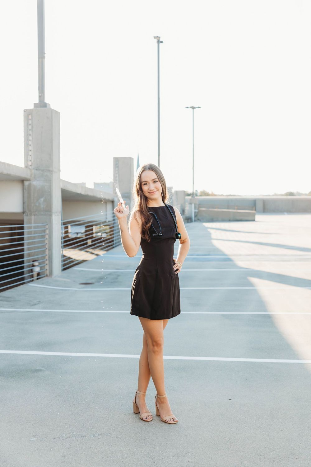 Young woman in black mini dress and heels posing in parking garage, pointing up with one finger.