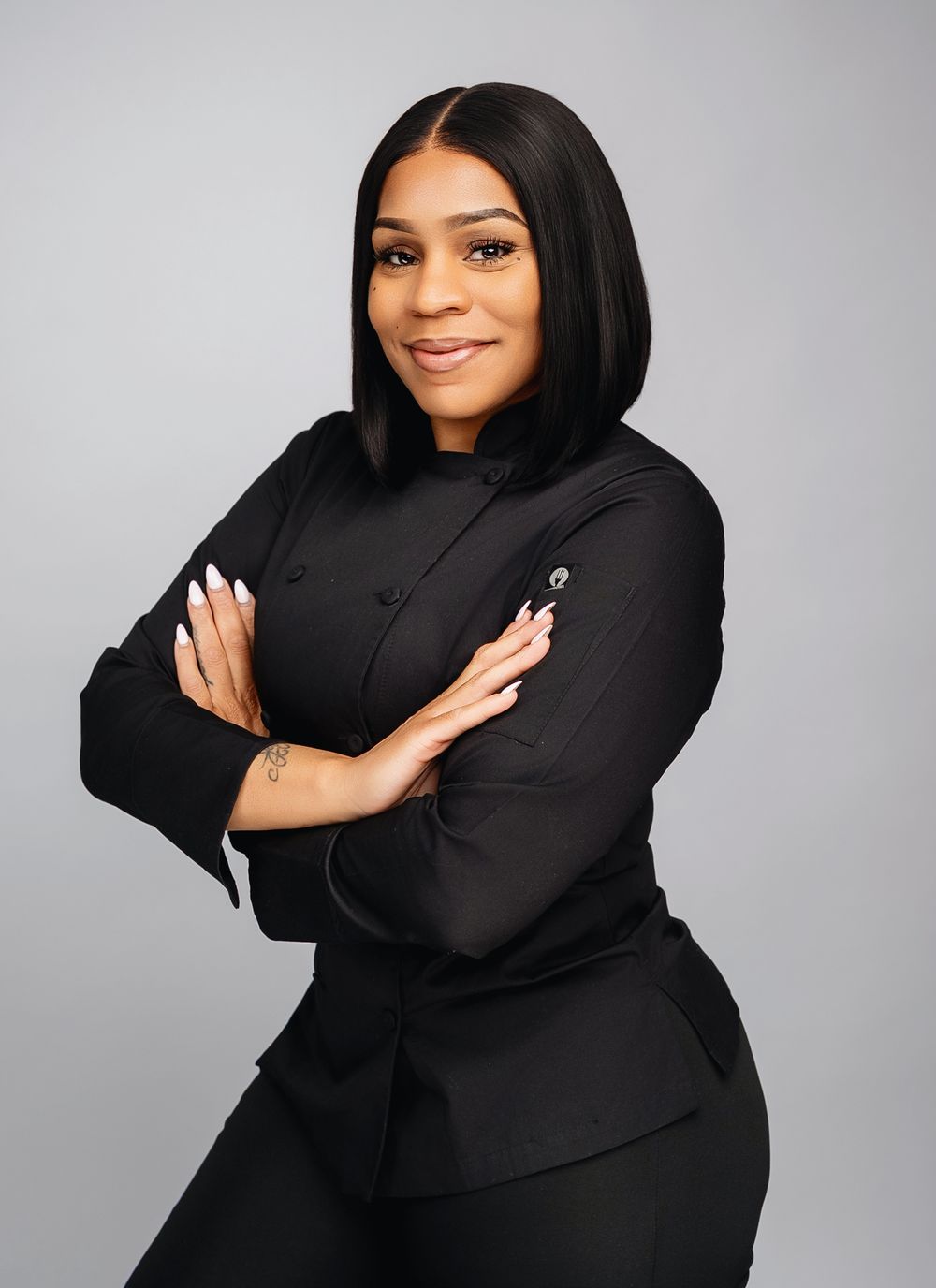 Professional portrait of a chef in a black coat standing confidently with arms crossed against a gray background.