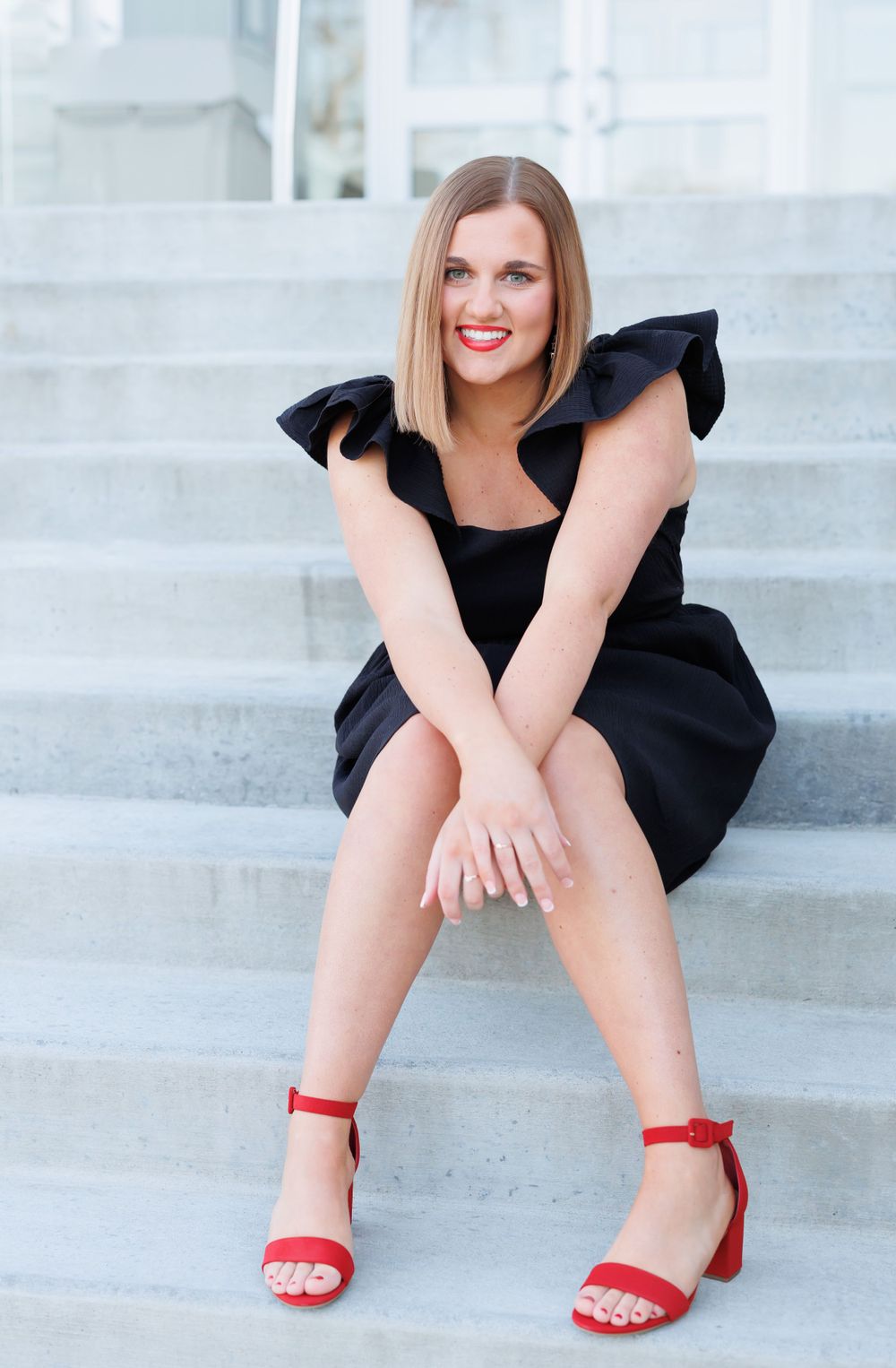 A woman in a black dress with ruffled shoulders sits on white steps wearing bright red sandals.