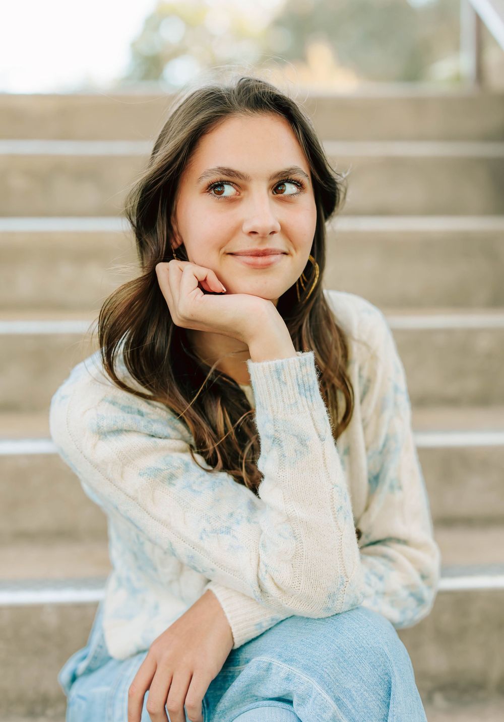 A person in a cozy cream sweater sits thoughtfully on concrete steps while looking off to the side.