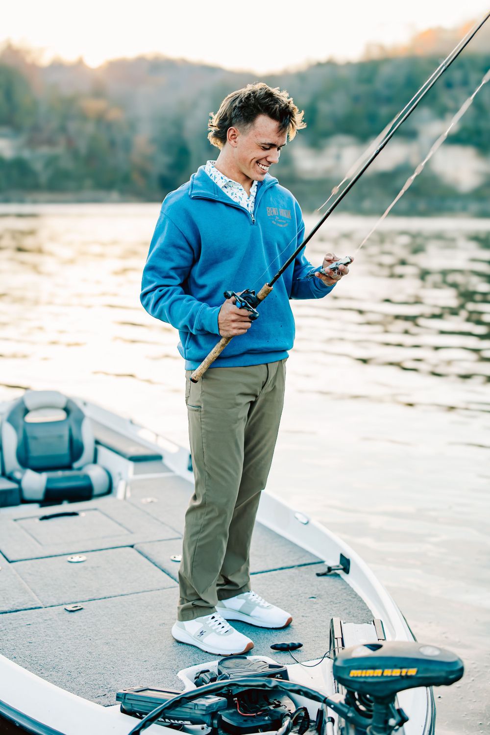 Angler in blue fleece hoodie checks smartphone while fishing from boat deck at sunset on calm lake waters.