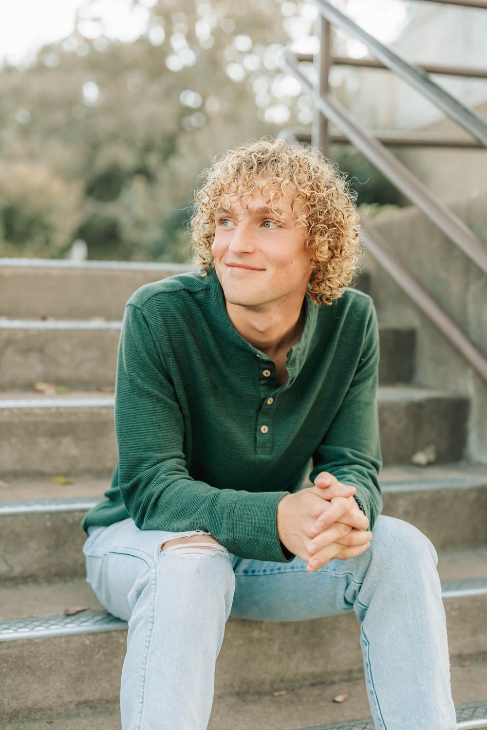 A person in a green henley shirt with curly blonde hair sits relaxed on concrete steps outside during golden hour.