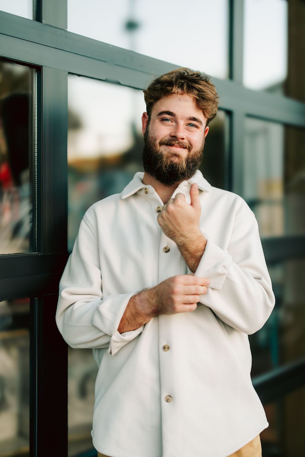 Smiling person in white button-down shirt fixing sleeve cuff while standing near glass window.