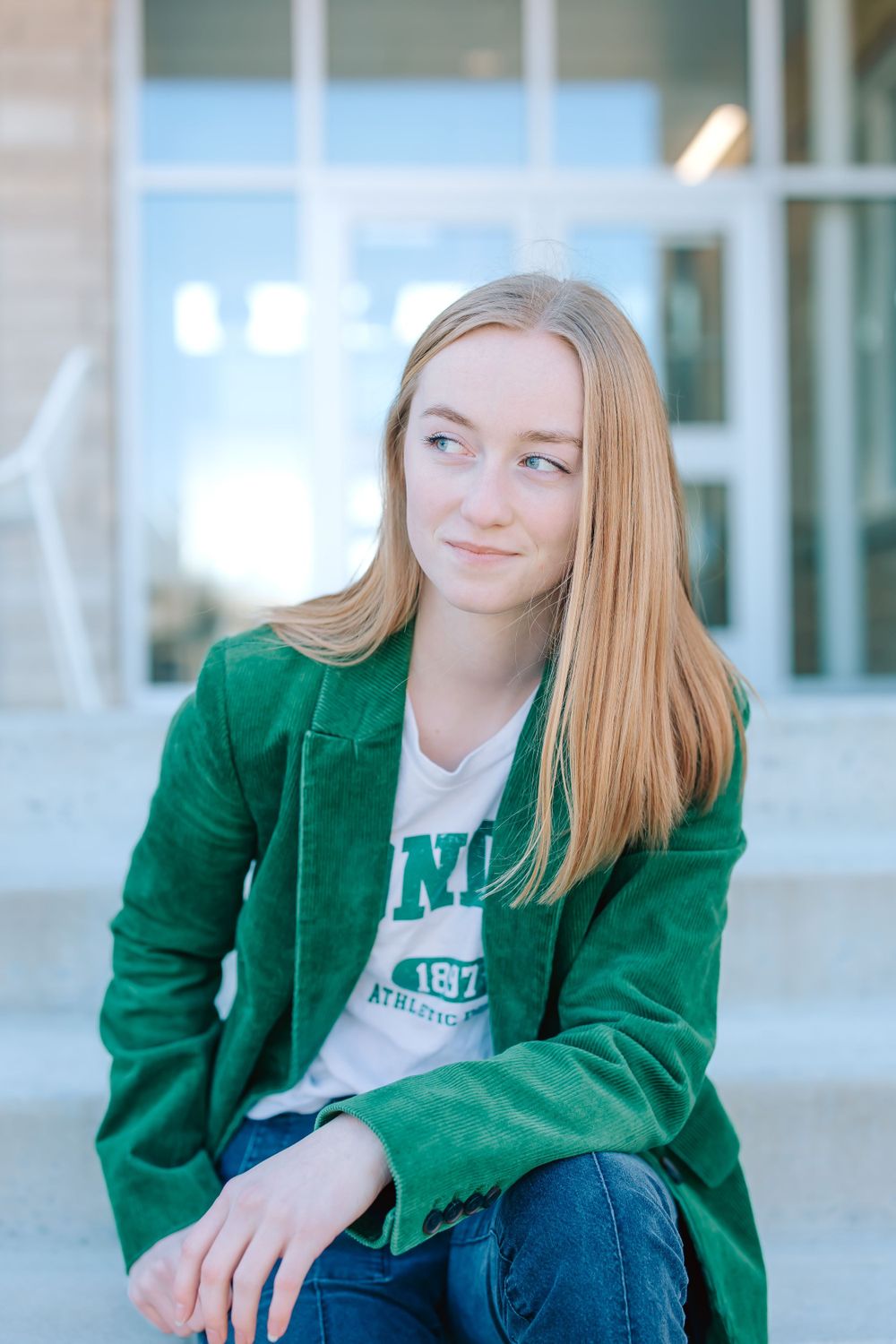 A young person with blonde hair wearing a green varsity jacket and jeans sits outdoors by a building.