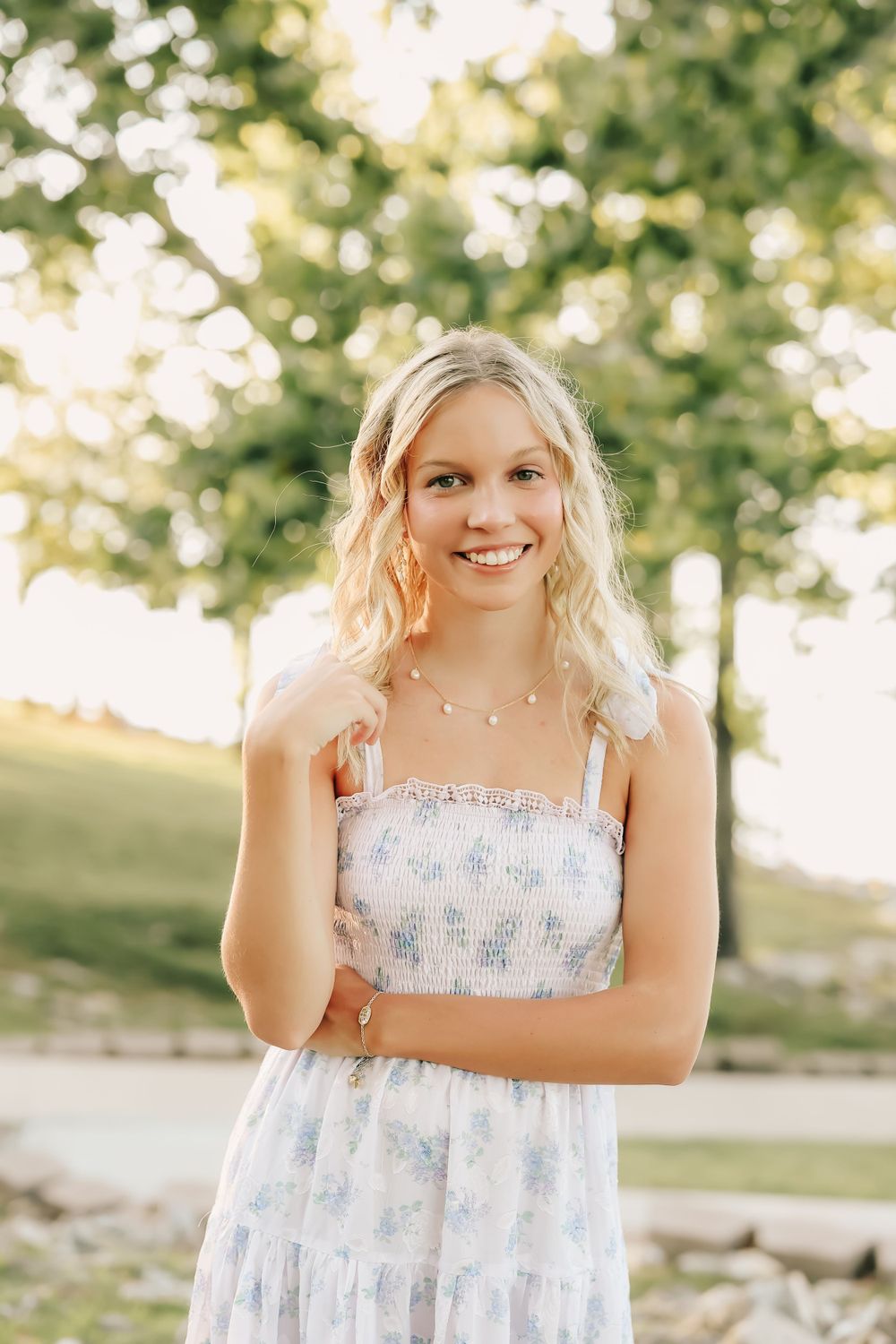 A young person in a white floral sundress smiles outdoors on a sunny summer day with bokeh trees in background.