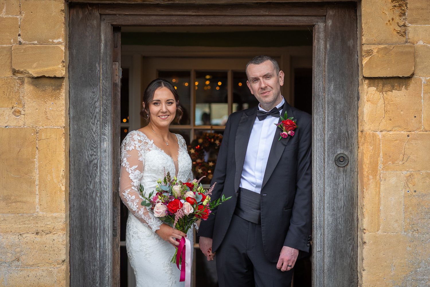 The bride and groom stand in a stone doorway on their wedding day at The Manor House Hotel, with the bride holding a red and white bouquet.