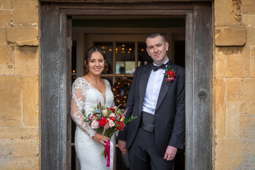 The bride and groom stand in a stone doorway on their wedding day at The Manor House Hotel, with the bride holding a red and white bouquet.