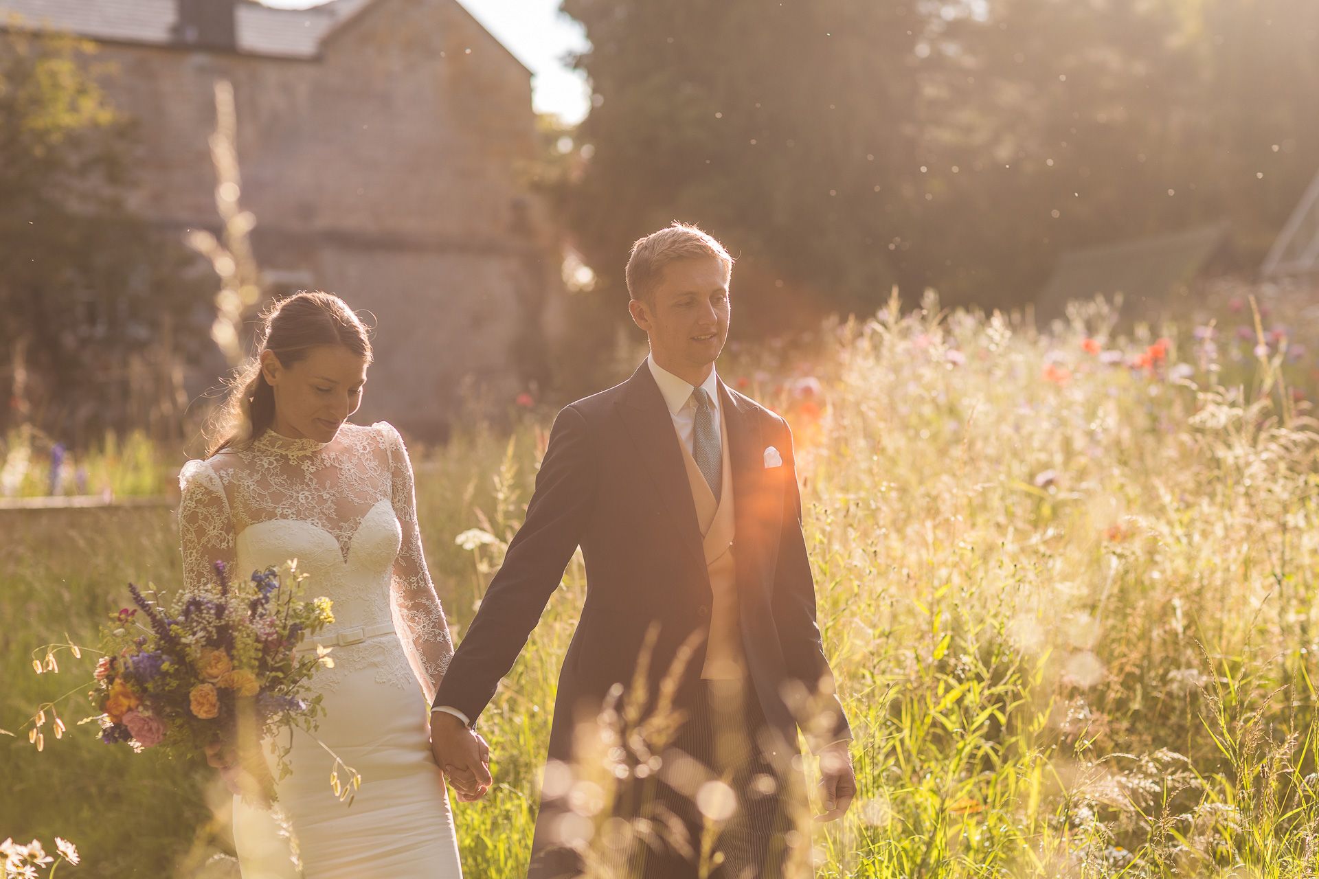 Bride and Groom walking through a sunlit meadow during golden hour with tall grass and flowers.