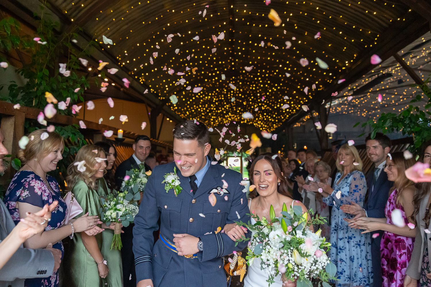 Wedding couple walks through a shower of flower petals and confetti under twinkly lights at Stone Barn as guests celebrate.