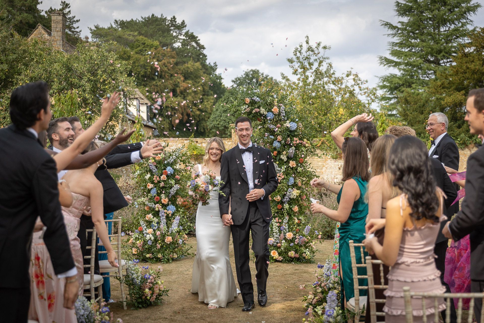 Wedding guests toss flower petals during an outdoor garden ceremony at Slaughters Manor House with floral arch and greenery.