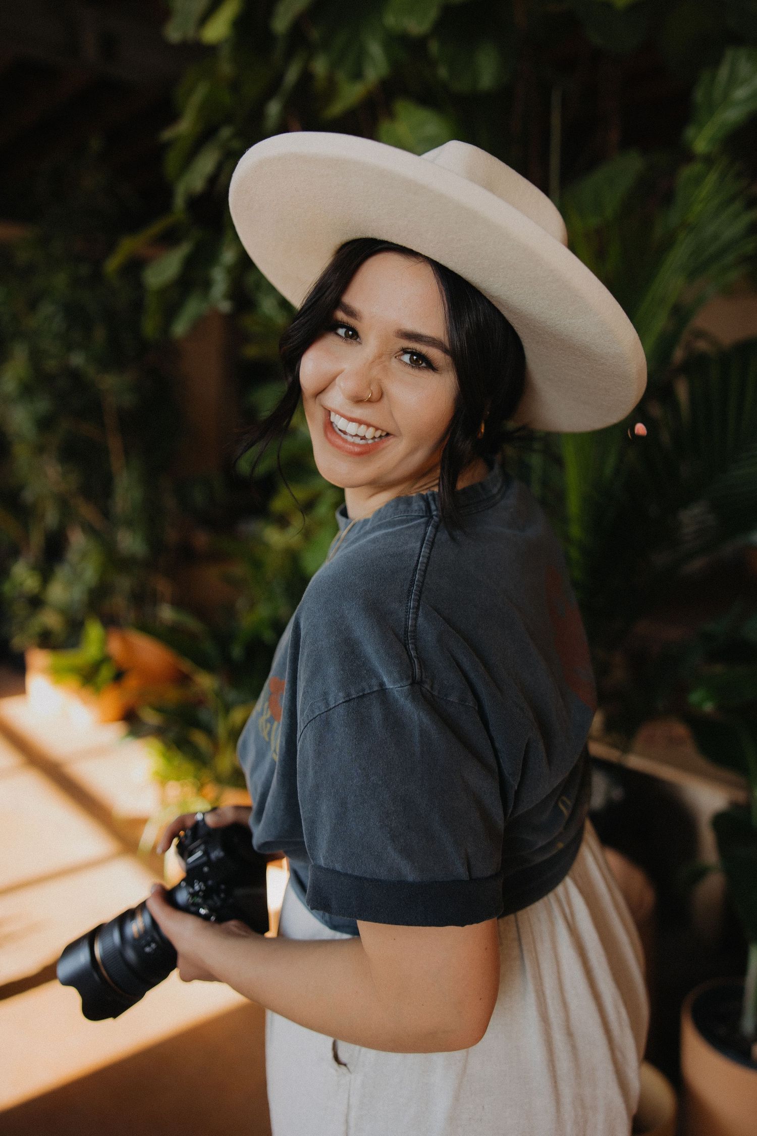 Photographer in a wide-brimmed hat holding camera while smiling in outdoor garden setting.