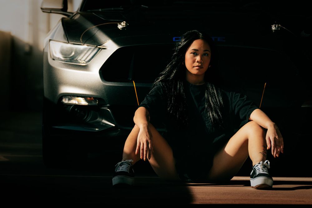 Stylish figure in black outfit sits on ground at night beside silver car in moody lighting.