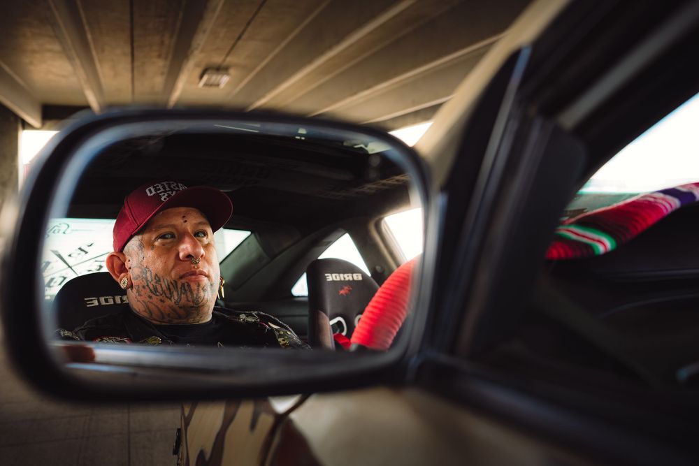 Car side mirror reflection showing interior view of vehicle with red cap and beard visible.