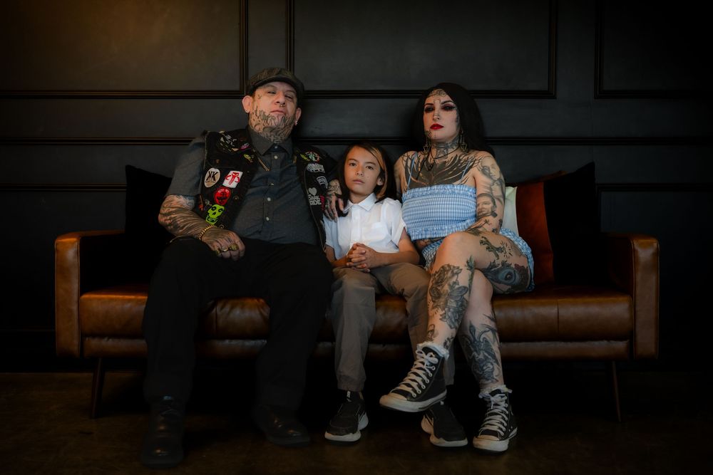A family adorned with tattoos sits together on a dark leather couch in a dimly lit setting.