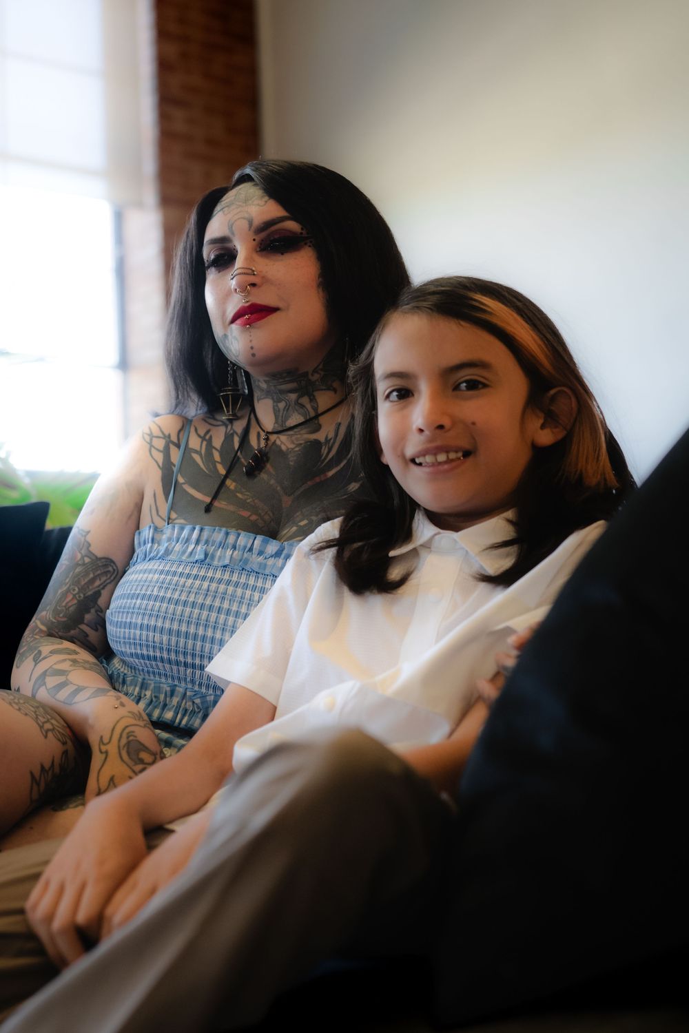 Two people sit together on a couch in a warmly lit room, one wearing a white shirt and the other in a checkered dress.