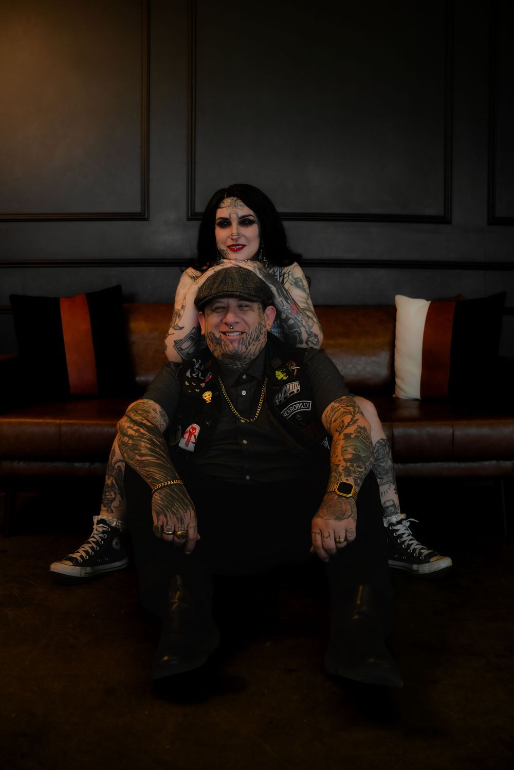 A tattooed couple sitting on a brown leather couch in a dark moody setting.