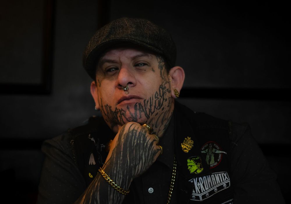 A person with facial tattoos wearing a flat cap and leather jacket lights up a match in dark lighting.
