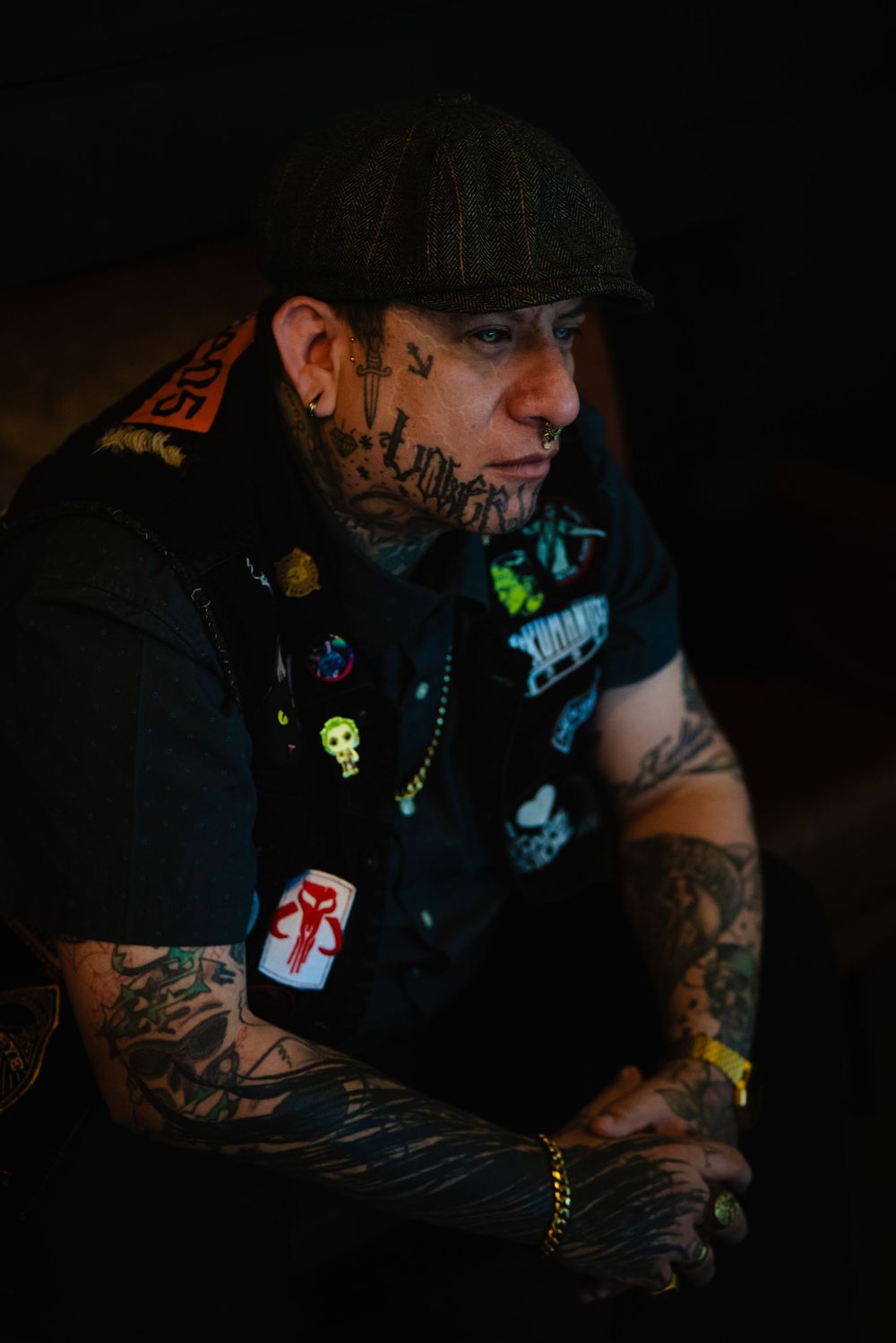 Person with tattoos wearing dark clothing and accessories in dimly lit setting.