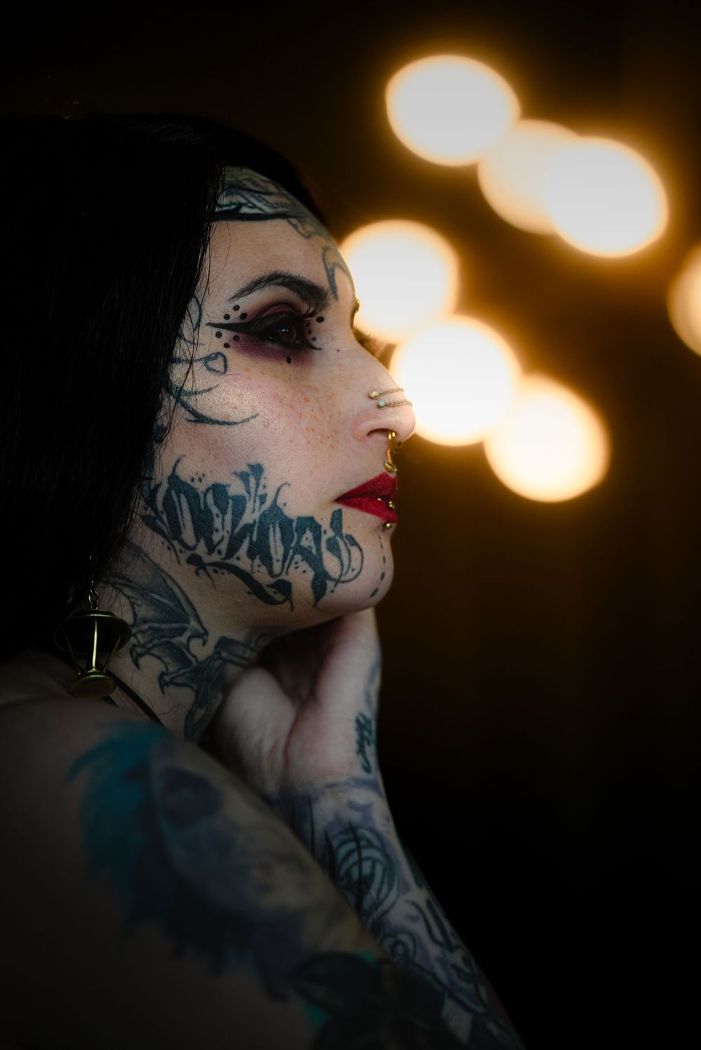 Artistic side profile portrait with dramatic bokeh lighting and neck tattoos against dark moody background.