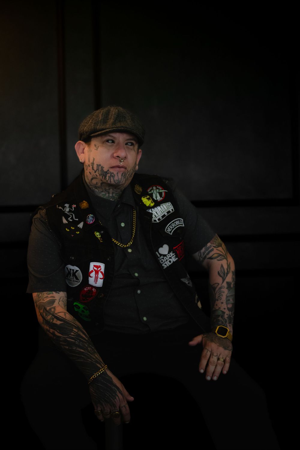Dark moody portrait showing a person in a black shirt adorned with patches, wearing a flat cap and displaying tattoos.