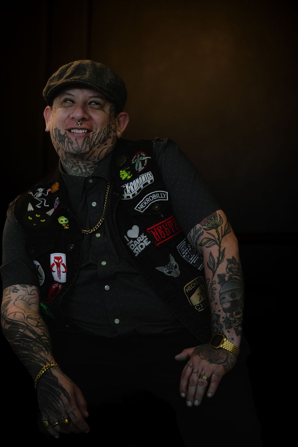 A heavily tattooed person in dark clothing and a flat cap smiles while wearing patches on their vest.