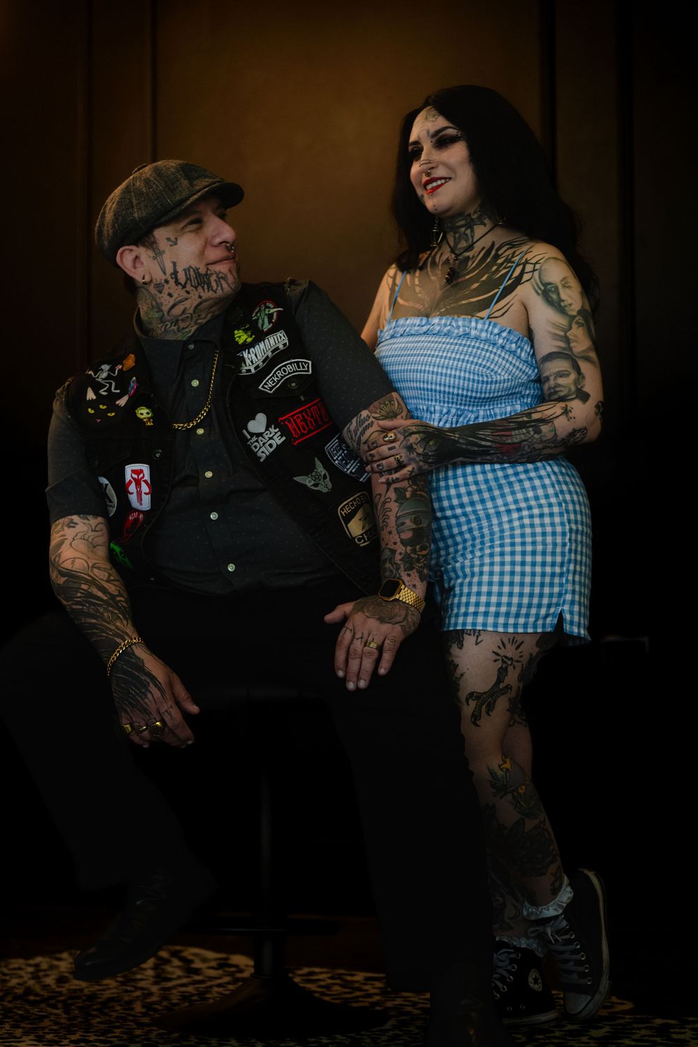 A heavily tattooed couple poses together in moody lighting, wearing contrasting outfits of black and gingham.