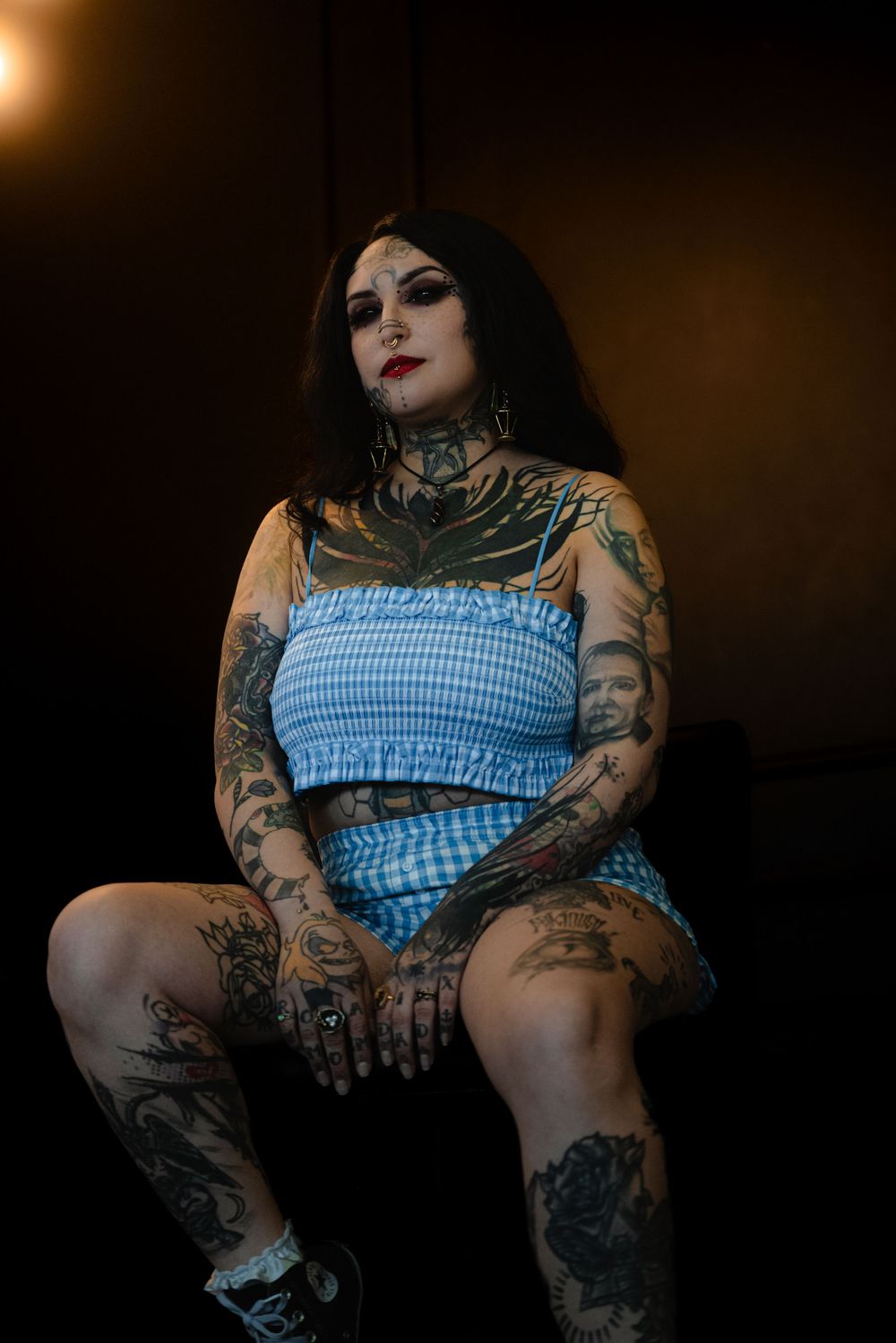 Woman in blue gingham dress with extensive tattoos poses in moody dark lighting against black background.