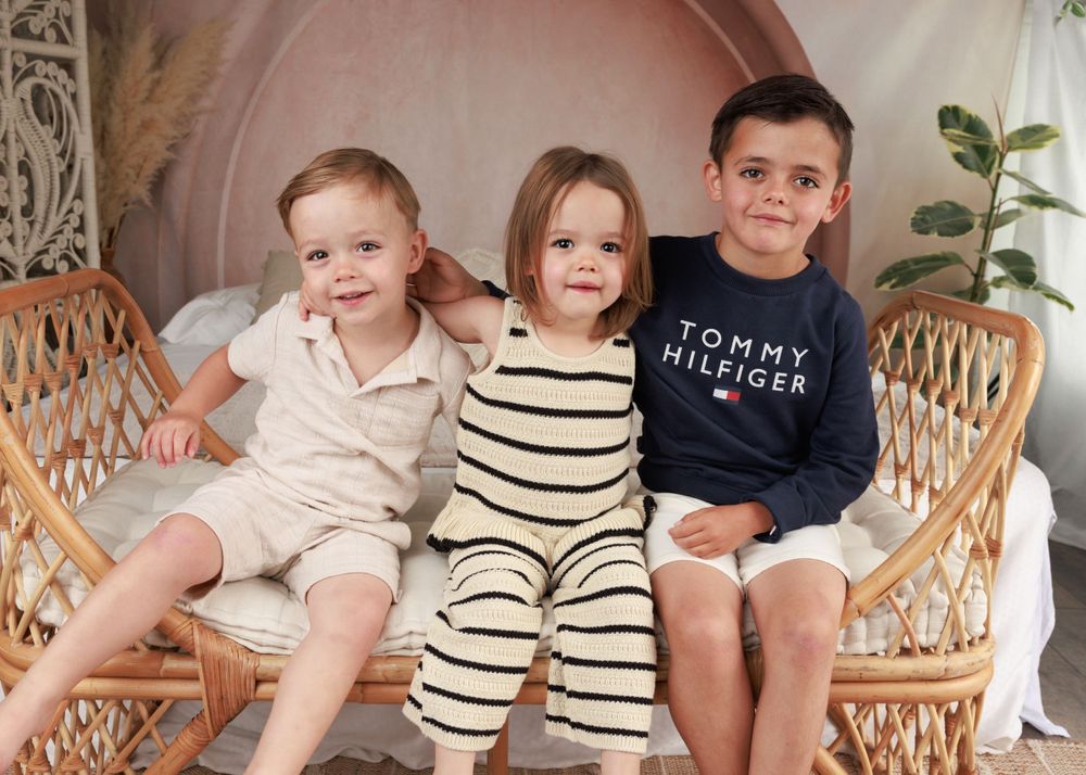 Three siblings posing in rattan chair against arched pink wall with plant.