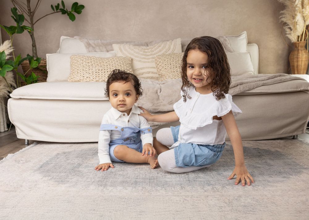 Young siblings sit together on a plush carpet in front of a white couch with decorative pillows and greenery.