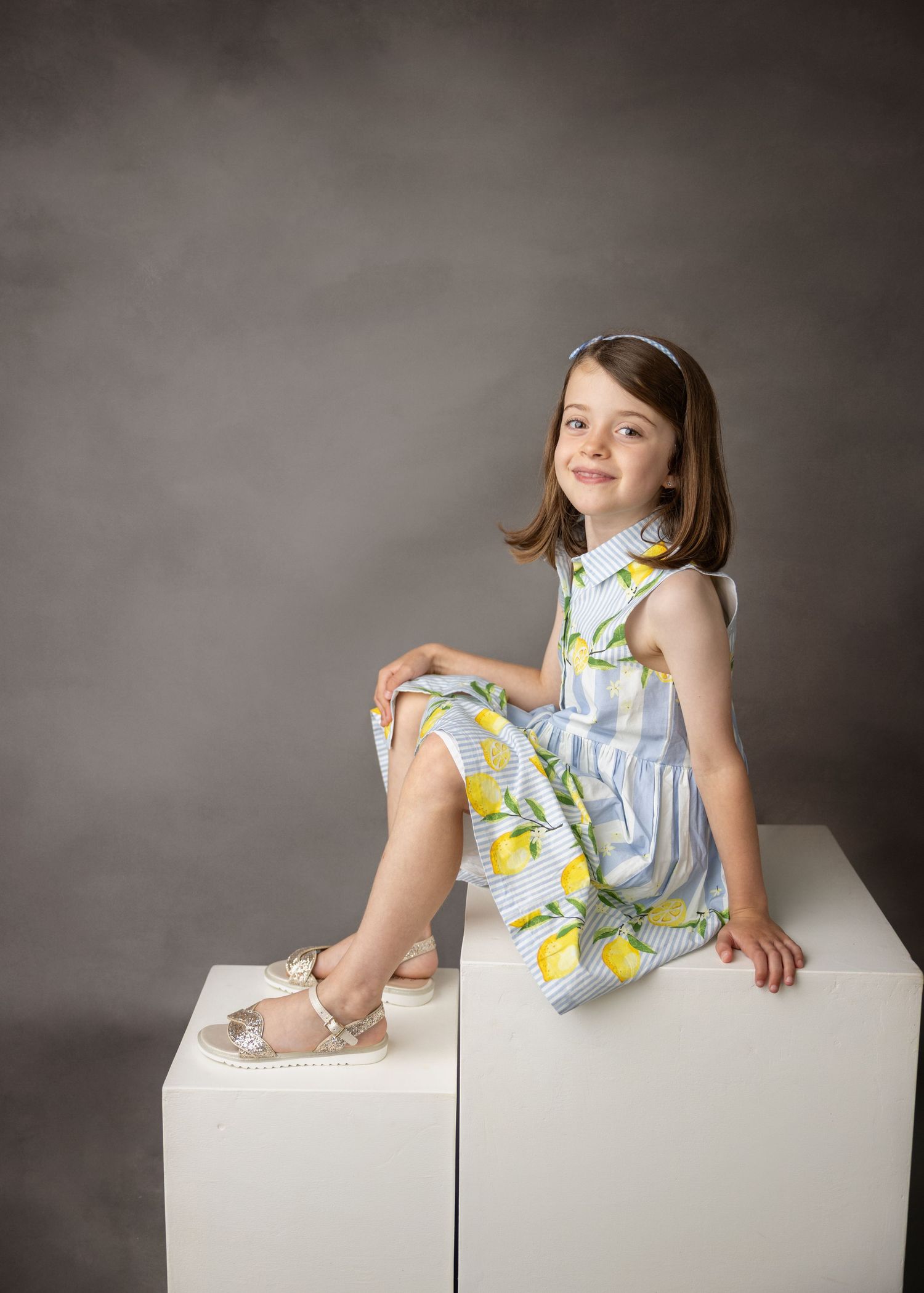 Smiling little girl in a blue striped lemon-print dress and silver sandals sits on white studio blocks against a gray backdrop.