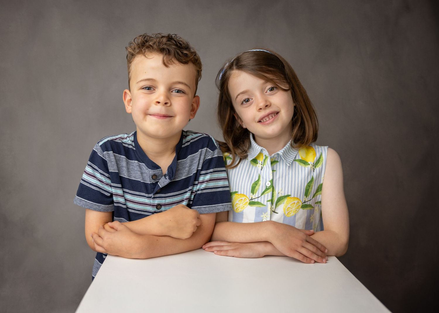 Smiling boy in a navy striped shirt and girl in a lemon print sleeveless top lean on a white table against a gray studio backdrop.