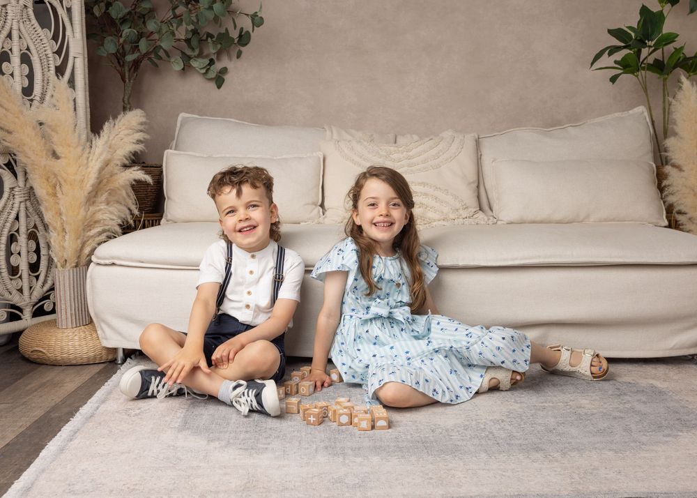 Two young children sit together on a light gray floor in front of a beige sofa with decorative pampas grass nearby.