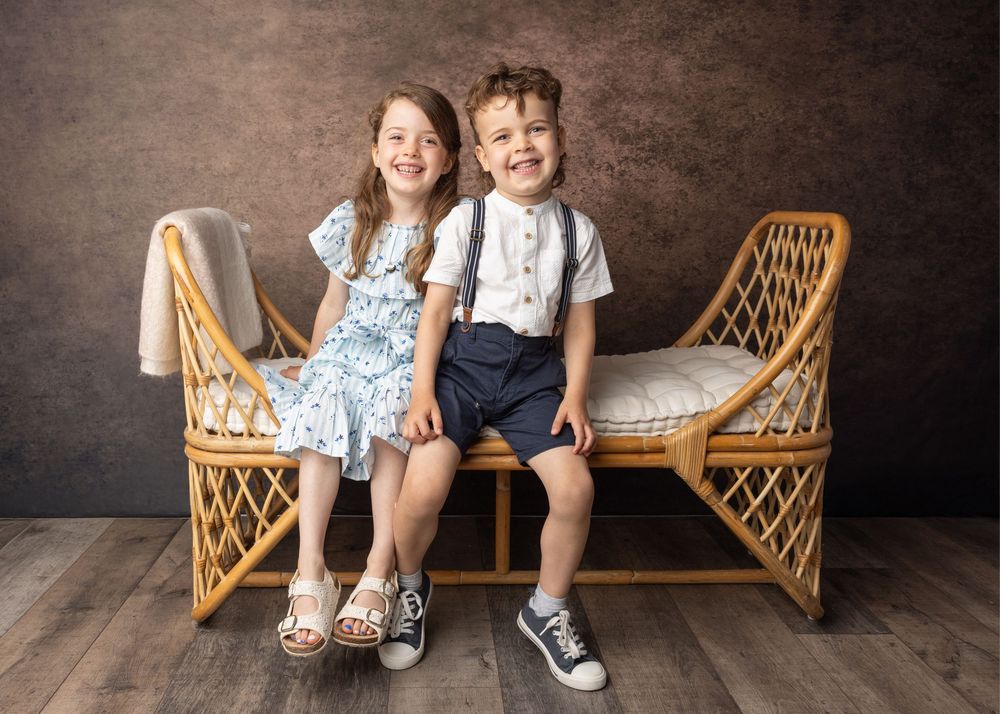 Young children sitting on a woven rattan bench pose playfully against a dark brown backdrop.