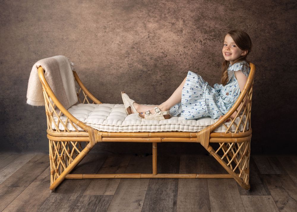 A young person lounges peacefully in a vintage wicker daybed with a light blanket in a studio setting.