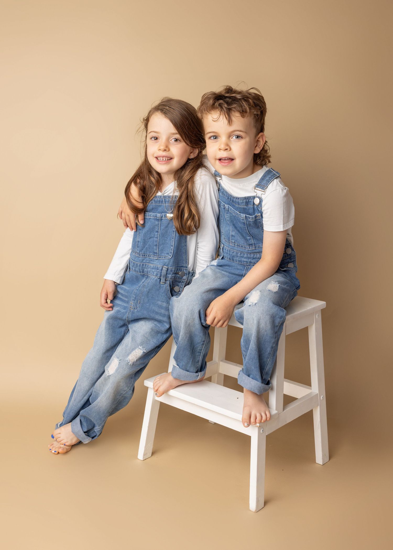 Two children in matching denim overalls sit together on a white wooden step stool against a beige backdrop.