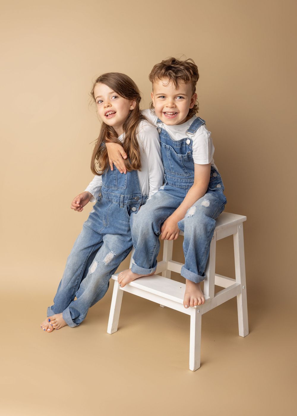 Two children in matching denim overalls laugh while sitting back-to-back on a white stepstool.
