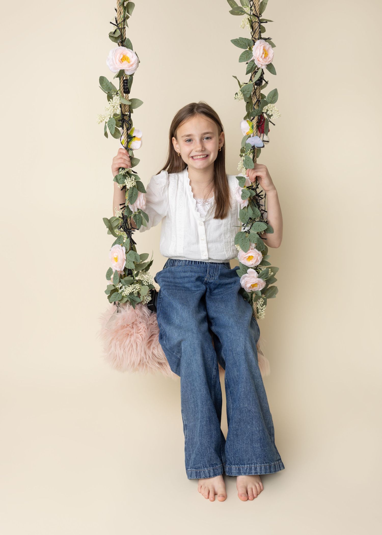 Young person on floral decorated swing with pink roses and greenery against neutral wall.
