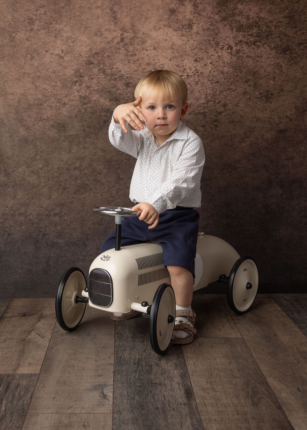 A small child in navy shorts and white shirt rides a vintage-style cream pedal car against a rustic brown backdrop.