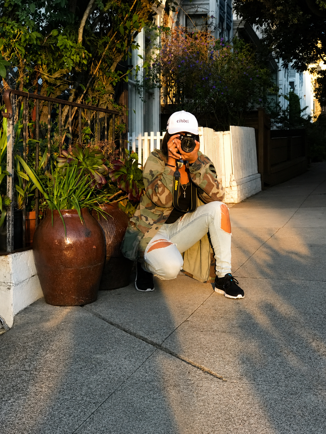 Photographer in camo jacket and white cap crouching on sidewalk, shooting with camera near large terracotta pot.