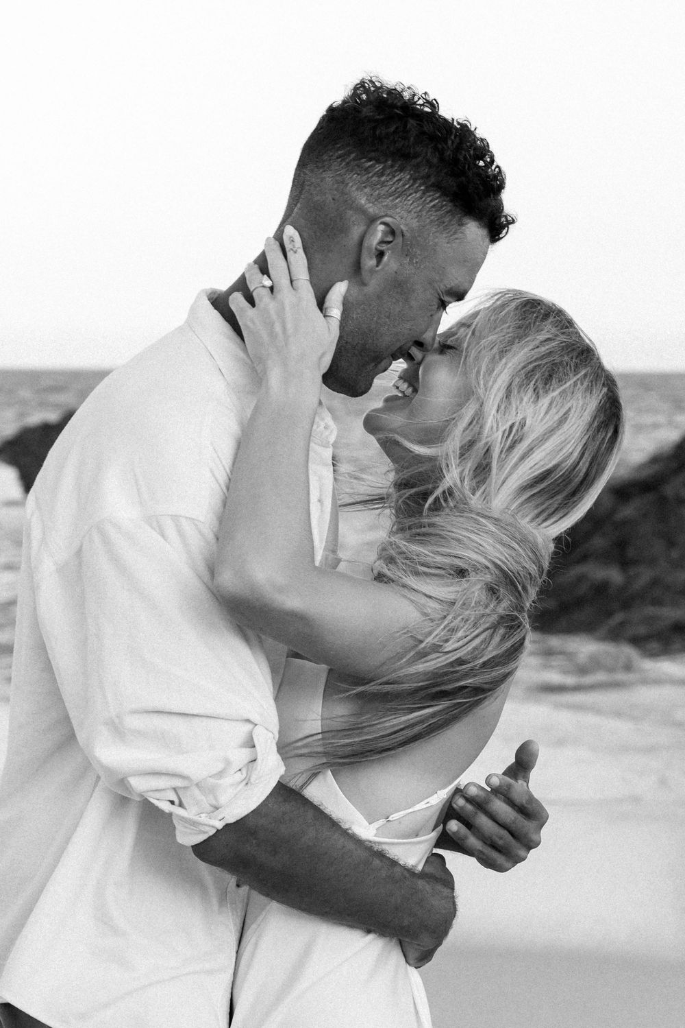Black and white close-up of a couple embracing and smiling at each other on a beach. The woman's hand rests on the man's