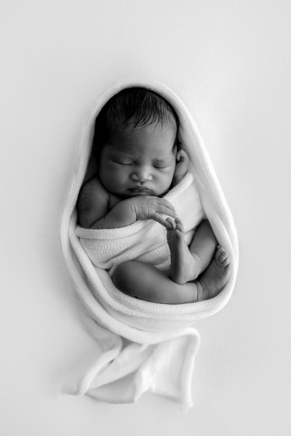 A series of black and white photos showing a sleeping newborn baby swaddled in white fabric in different positions.