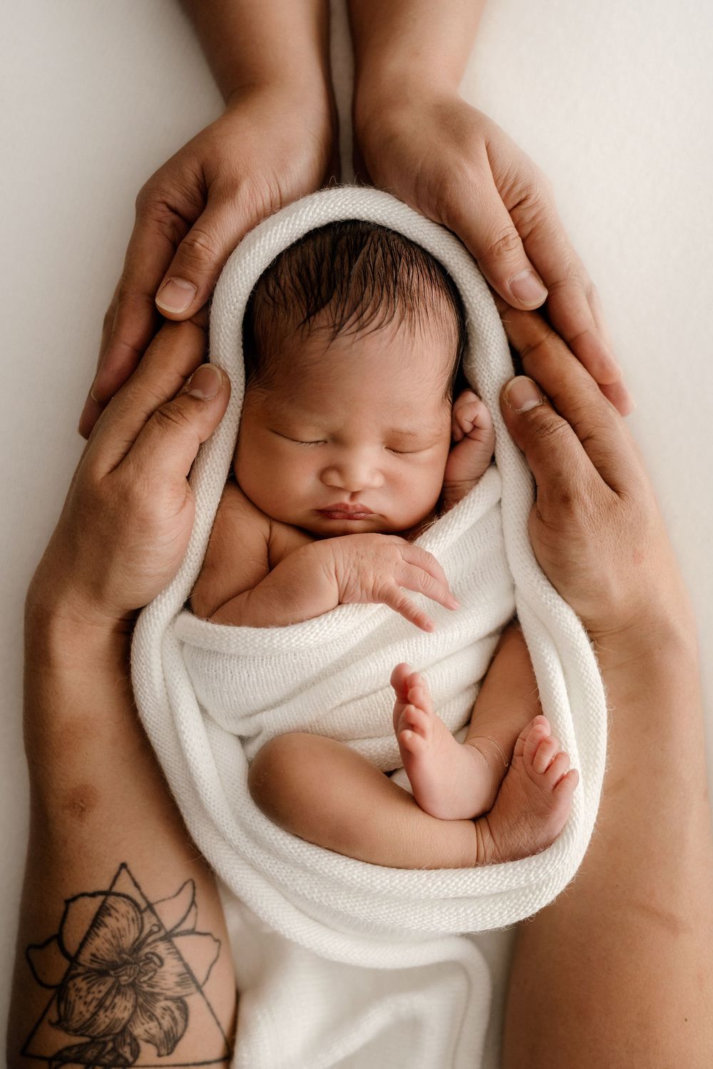 A sleeping newborn baby wrapped in white fabric is gently cradled by tattooed hands in a series of tender poses.